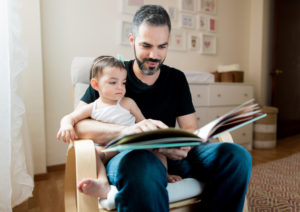 Un padre sentado en la silla con un bebé leyendo un libro. Hay pocos programas de educación disponibles para ayudar a papás.