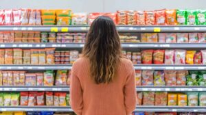 Mujer recorriendo el pasillo de snacks en un supermercado. Los estantes están llenos de diversos alimentos envasados. Crédito de la imagen: Adobe Stock, creada con IA.