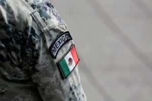 Primer plano del hombro de un uniforme militar con camuflaje pixelado gris, mostrando un parche bordado con la palabra 'MEXICO' y la bandera mexicana.